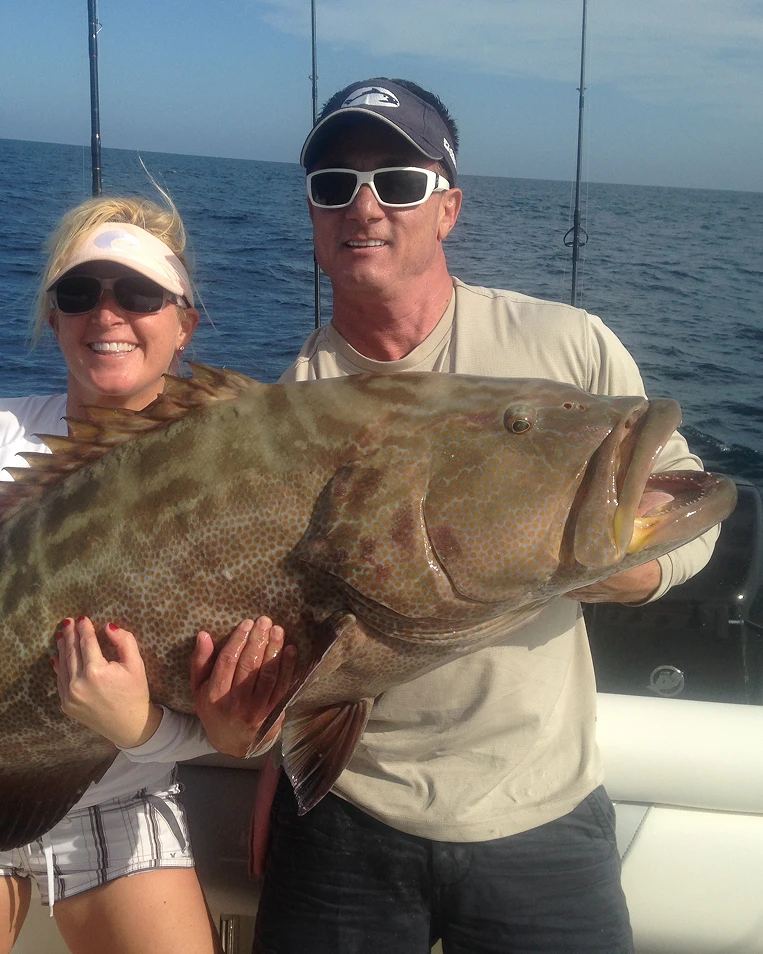 Two people on a boat holding a very large fish with the ocean in the background.