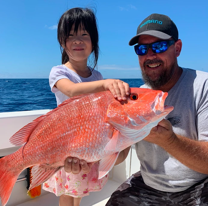 A man and a girl on a boat proudly holding a large red snapper fish against a clear blue ocean and sky backdrop.