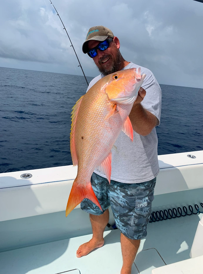 Man on a boat holding a large orange and pink fish with the ocean and a cloudy sky in the background.