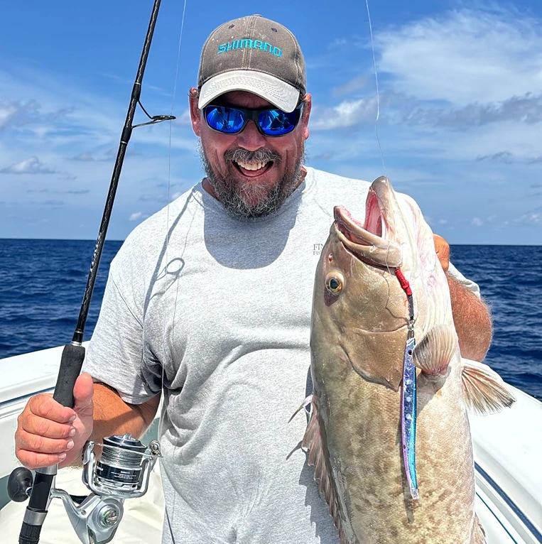 Man on a boat holding a fishing rod in one hand and a large caught fish in the other under a clear blue sky.