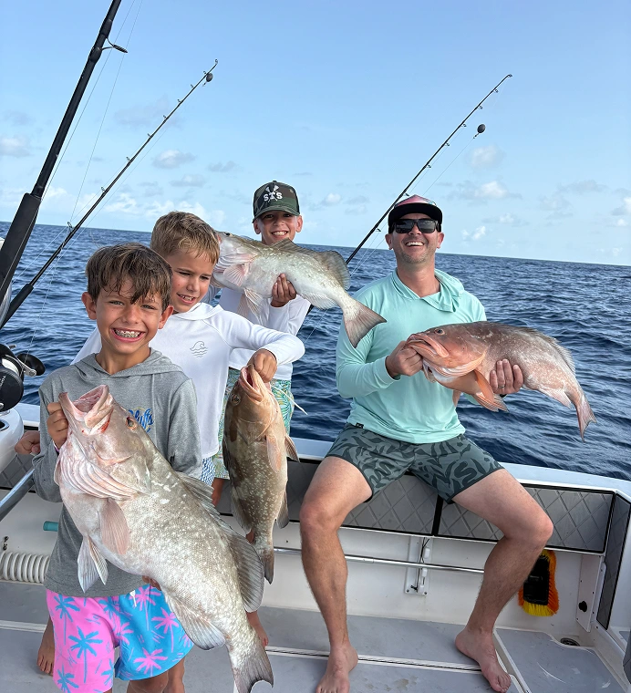 Four people on a boat holding large fish, with fishing rods and the ocean visible in the background.