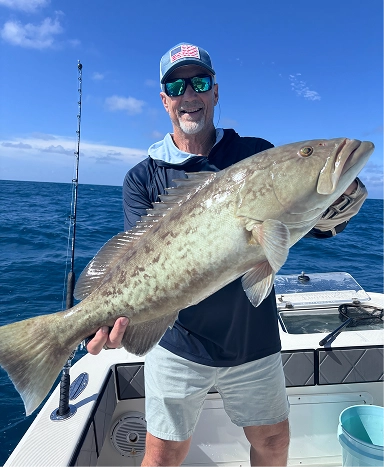 A person on a boat holds a large fish, with a blue sky and ocean in the background. Fishing gear is visible on the boat.