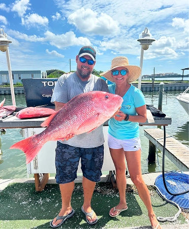 A person holds a large red snapper fish while standing on a dock near a marina, with boats and blue skies in the background.