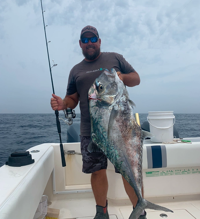 Man on a boat holding a large fish and a fishing rod with the ocean and cloudy sky in the background.