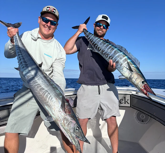 Two men on a boat holding large caught fish with ocean and clear blue sky in the background.