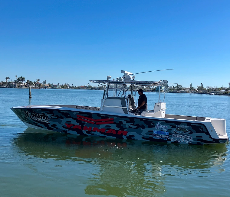 Fishing boat with camouflage design and sponsor logos floating on calm water under clear blue sky.