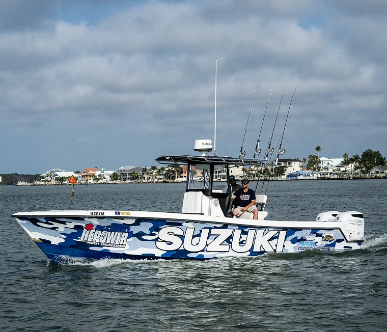 Fishing boat with blue camouflage design and large Suzuki logo cruising near a coastal town on a cloudy day.