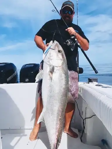 Person standing on a boat holding a large silver fish with a fishing rod, under a clear blue sky.