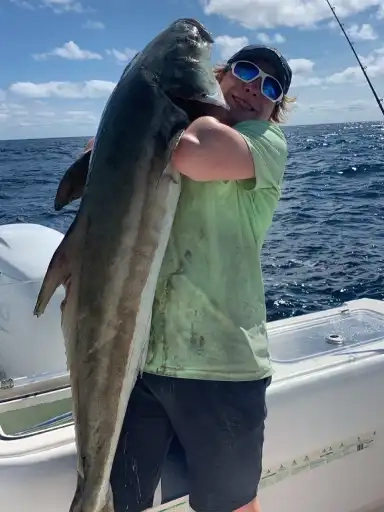 A person on a boat holds a large fish with ocean waves in the background. They're smiling, wearing sunglasses and a cap, conveying excitement and achievement.