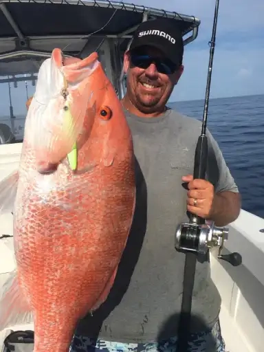 Man on a boat smiling, proudly holding a large red snapper and a fishing rod. He's wearing a cap and sunglasses, with the ocean in the background.
