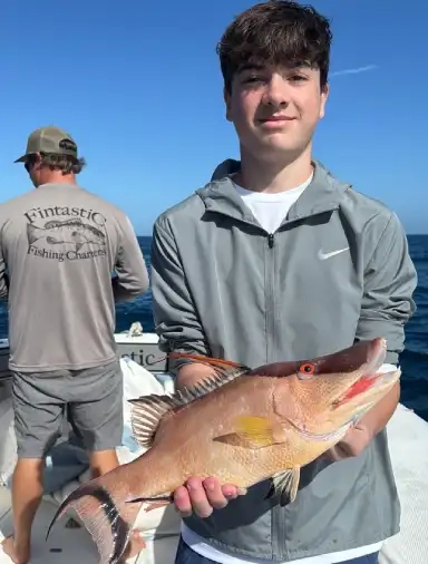 A young man on a boat holding a large fish proudly. Another person in the background wears a fishing charter shirt. Clear blue sky and ocean view.