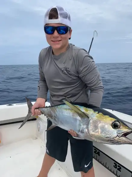 A person in a gray shirt and cap, wearing sunglasses, smiles while holding a large fish on a boat, with a calm sea and cloudy sky in the background.