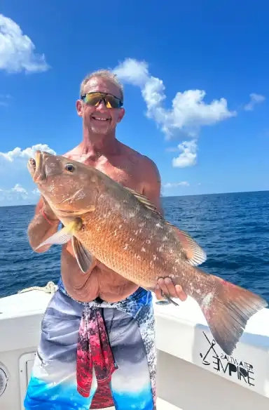A man stands on a boat holding a large fish, smiling under a bright blue sky with fluffy clouds. The ocean is calm, emphasizing a joyful fishing moment.
