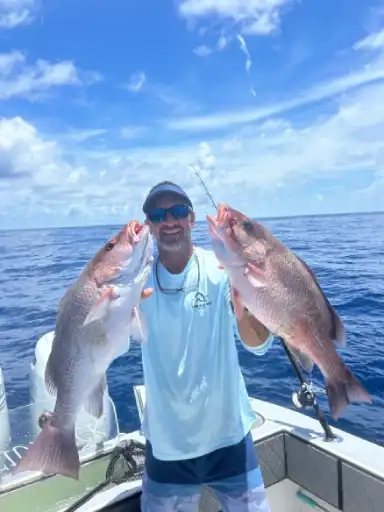 A smiling man on a boat proudly holds two large fish, with a clear blue sky and ocean backdrop. He wears sunglasses and a light blue shirt, conveying a sense of accomplishment.