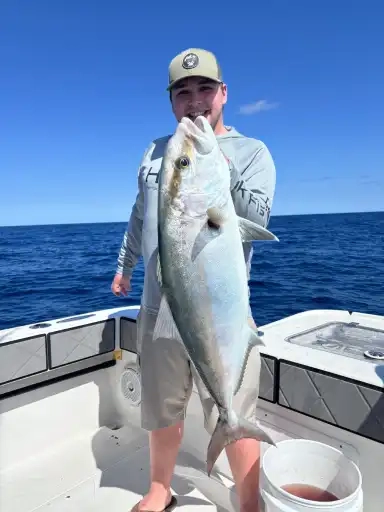 A person on a boat holds a large fish with both hands, smiling under a clear blue sky. The ocean stretches to the horizon, creating a serene backdrop.