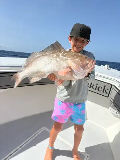 Young boy on a boat, smiling and holding a large fish. He's wearing a cap, light gray top, and bright pink shorts with blue palm tree patterns.