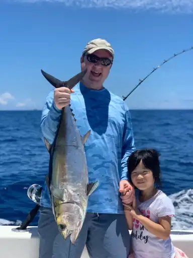 A man wearing sunglasses and a cap proudly holds a large fish on a boat. A smiling little girl stands beside him. The ocean and clear blue sky are in the background.