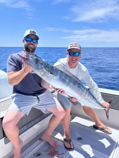 Two men on a boat holding a large fish, both smiling under a clear blue sky. The ocean is calm, and they appear excited and proud of their catch.