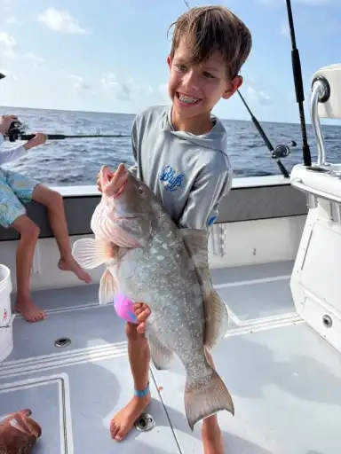 A smiling boy on a boat proudly holds a large fish, standing barefoot on the deck with ocean in the background, conveying excitement and achievement.