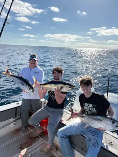 Three people on a boat holding large fish, with water and blue sky in the background.