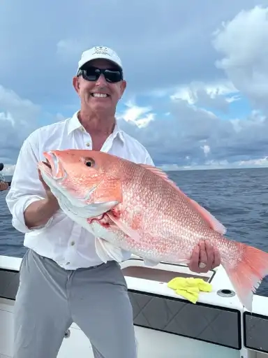 Person on a boat holding a large red snapper fish with the ocean and cloudy sky in the background.