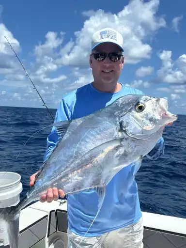 Person on a boat holding a large silver fish against a backdrop of blue ocean and partly cloudy sky.