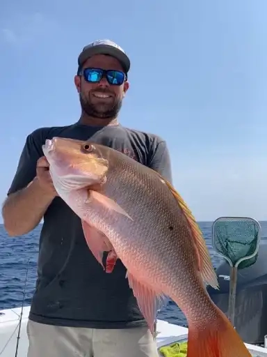 Person on a boat holding a large red snapper fish with the ocean and clear sky in the background.