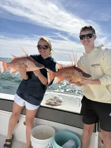Two people on a boat holding large fish with the ocean and blue sky in the background.