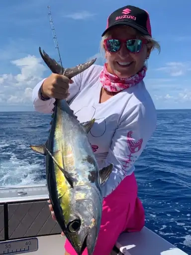 Person on a boat holding a large yellowfin tuna, dressed in pink and white fishing gear with sunglasses, ocean and blue sky in the background.