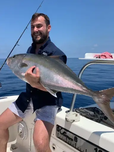 Man on a boat holding a large fish with a broad smile. The ocean is calm and blue under a clear sky, conveying a sense of accomplishment and joy.