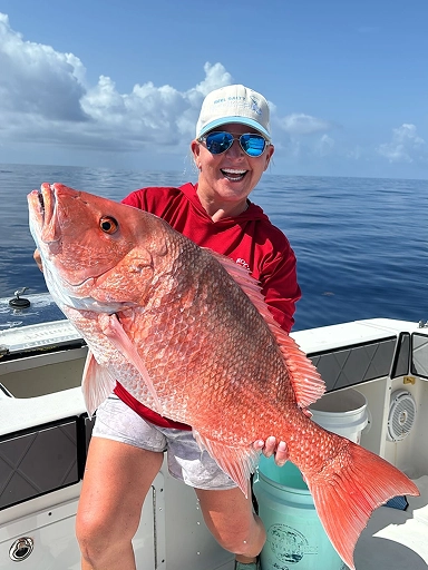 A person holds a large red snapper while standing on a boat, with calm sea and blue sky in the background.
