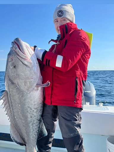 A person in a red jacket holds a large fish while standing on a boat in the ocean, with clear skies in the background.