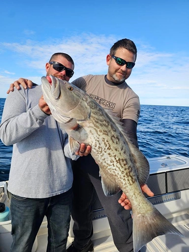 Two individuals on a boat hold a large, spotted fish against a backdrop of clear blue skies and water.
