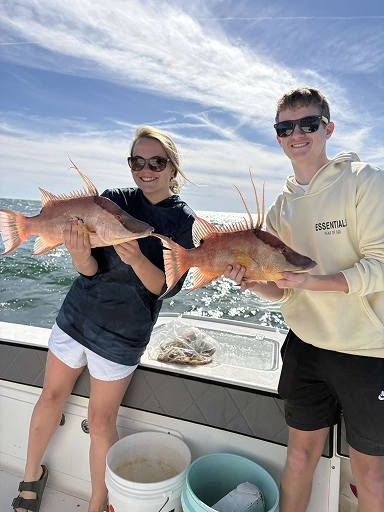 Two individuals on a boat hold up large pink fish, with water and blue skies in the background. Fishing gear is visible on the deck.