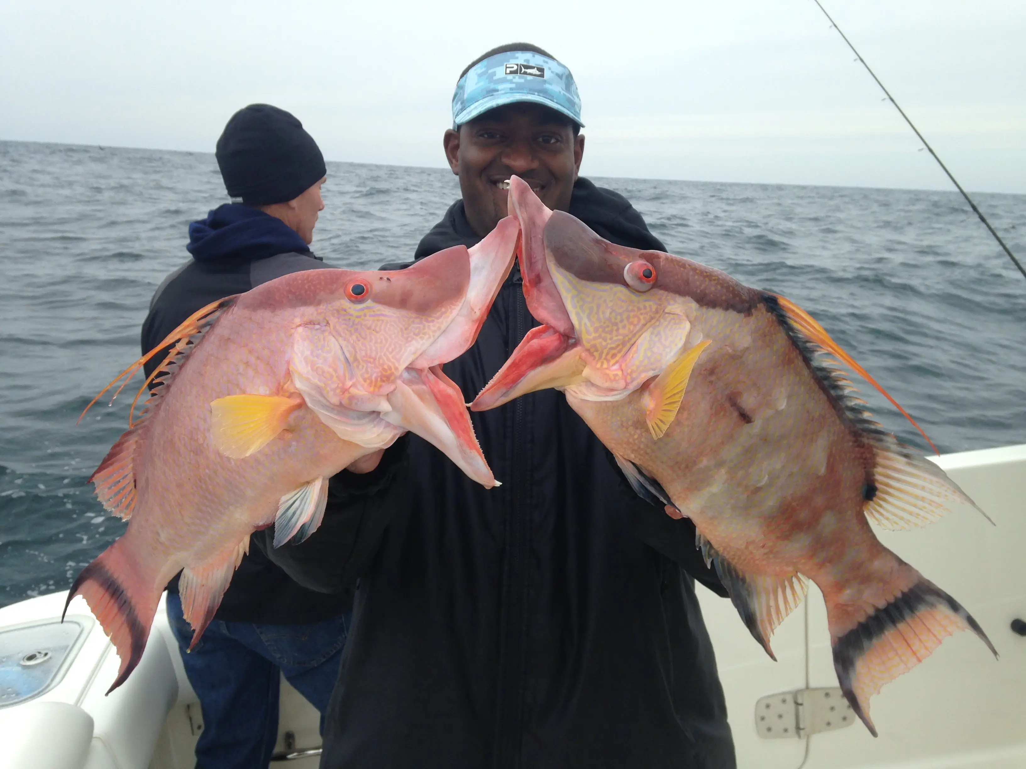 A smiling fisherman on a boat holding up two colorful Hogfish, with their unique snouts touching, against a backdrop of the grey open ocean.