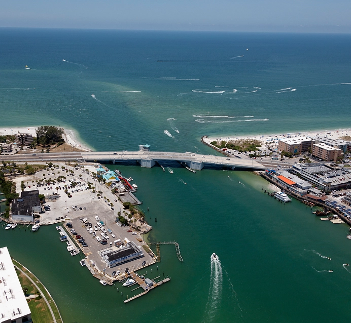 Aerial view of a coastal bridge over green water with boats, a marina, sandy beaches, and buildings under a clear blue sky.