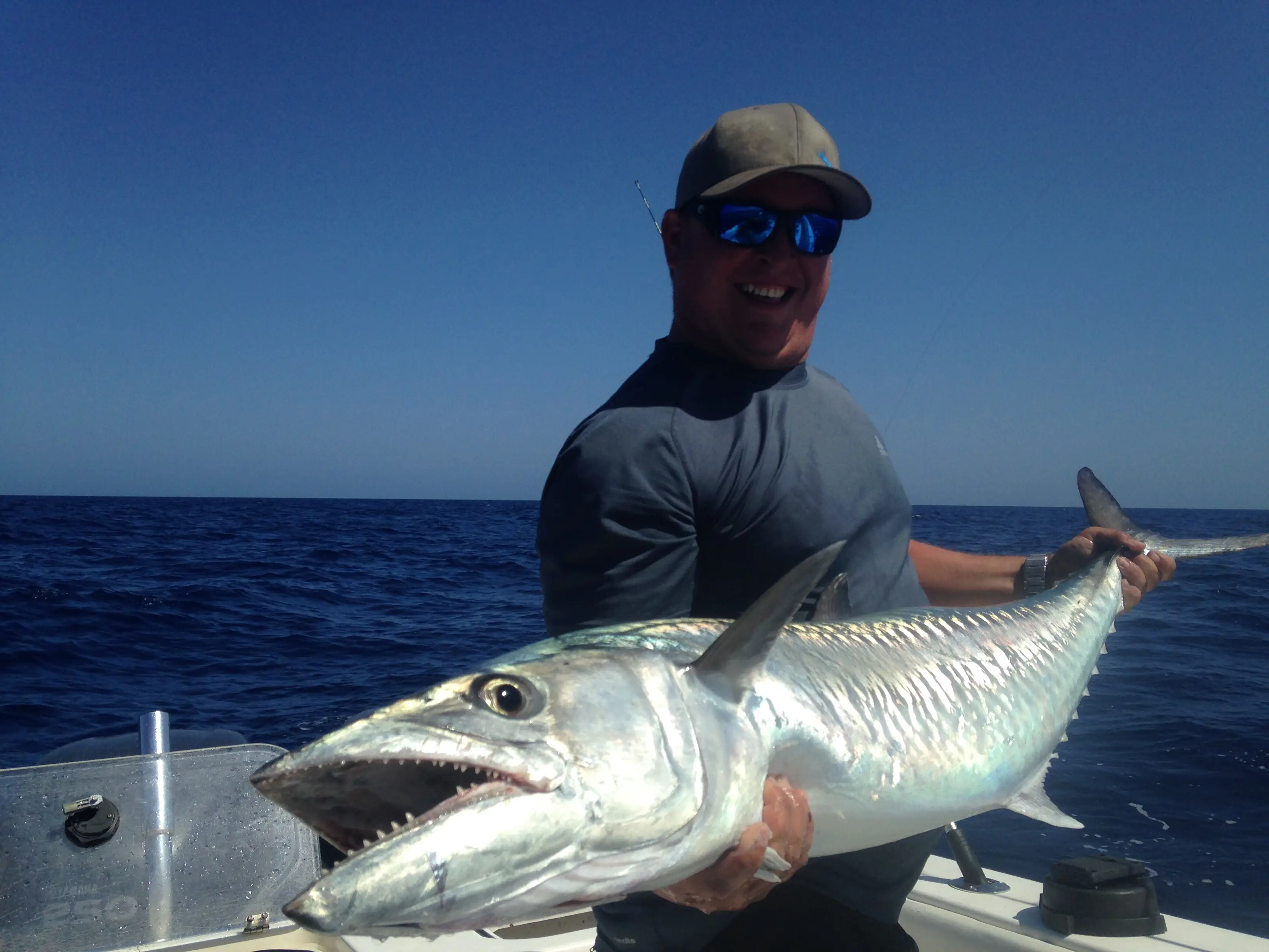 A smiling fisherman on a boat holding a large, silver King Mackerel with its mouth open, showing sharp teeth, against a clear blue sky and ocean background.