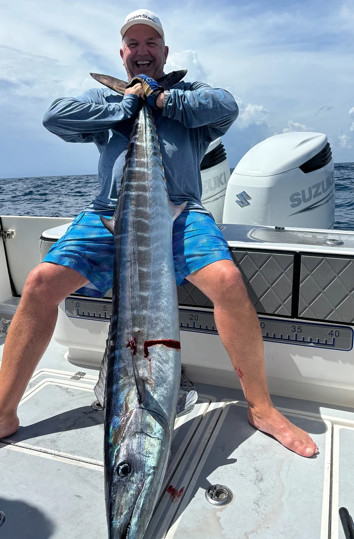 A person holds a large, shiny fish on a boat, with visible ocean and cloudy sky in the background. Blood is noted on the fish's body.