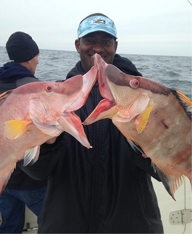 A person holds two large fish, showcasing their catch while on a boat against a cloudy ocean backdrop.