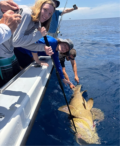 A group of people on a boat assist in reeling in a large fish, surrounded by deep blue ocean and clear skies.