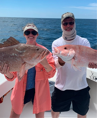 Two people on a boat proudly hold large fish: a grouper and a snapper, with blue ocean and sky in the background.