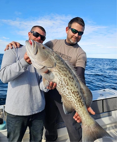Two individuals proudly hold a large fish on a boat in open water, with a clear blue sky in the background.