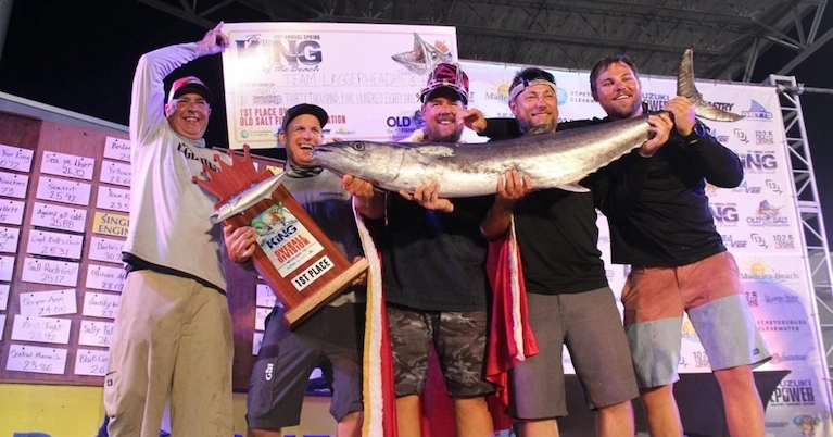 Five people holding a large fish and a 1st place trophy at a fishing competition on a stage with sponsor banners.