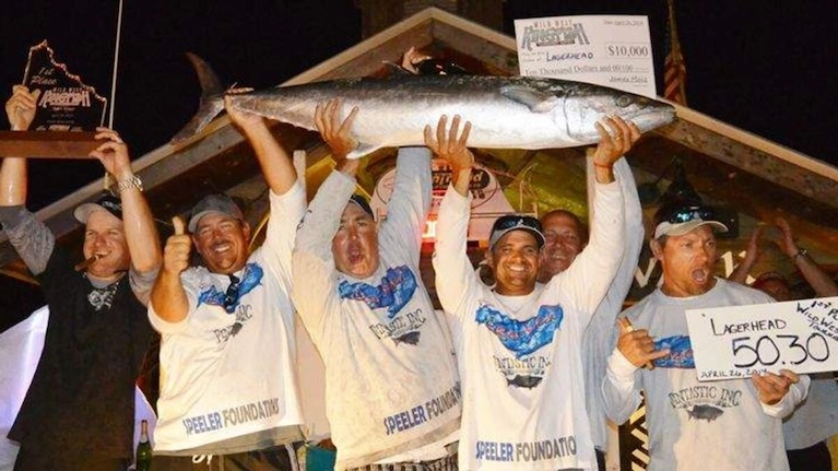Group of people holding a large fish and oversized checks and trophies at a night fishing competition celebration.