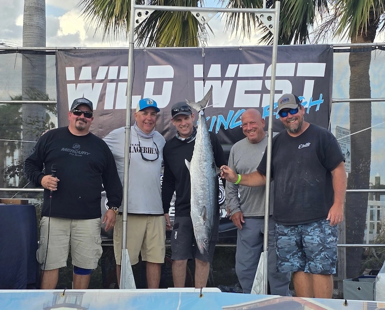Five men pose with a large fish hanging from a display frame at a Wild West Fishing Fest event.