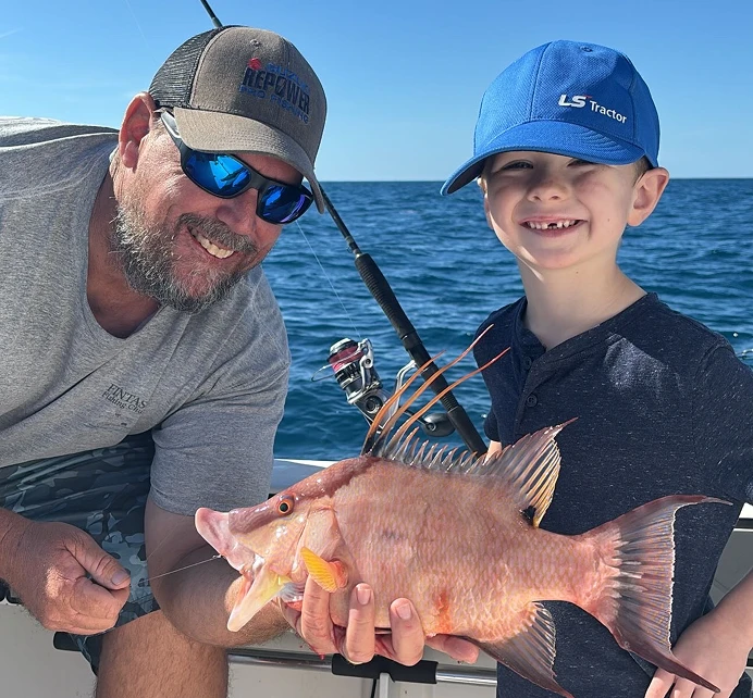 An adult and child proudly display a large pink fish on a boat, with the ocean visible in the background under a sunny sky.